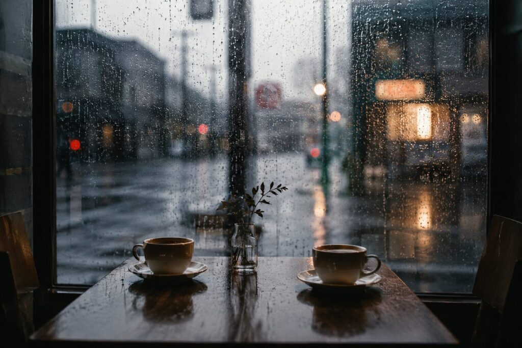 雨粒が流れる喫茶店の窓際。向かい合う二つのコーヒーカップの向こうに、ぼやけた雨の街並みが広がる静かな午後の風景。